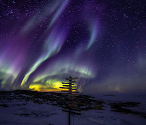 Purple yellow and green aurora australis across a night sky with a signpost in the foreground