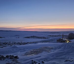 A building in the distance with lights on, surrounded by snow, ice and rocks, under blue sky at sunset