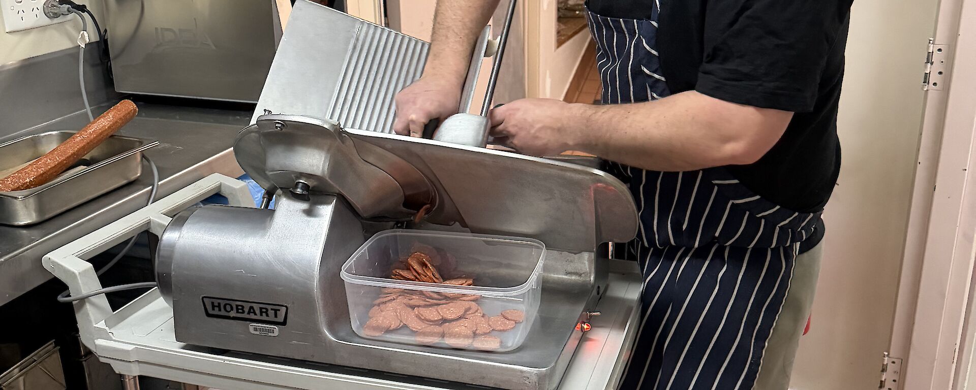 An expeditioner in the kitchen, slicing pepperoni with a large meat-slicing machine