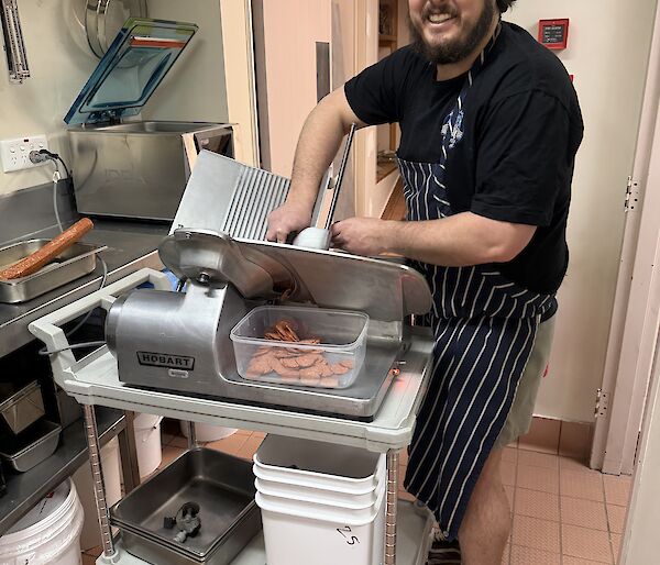 An expeditioner in the kitchen, slicing pepperoni with a large meat-slicing machine