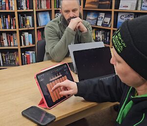 A person in a beanie on a computer in a library with someone watching on