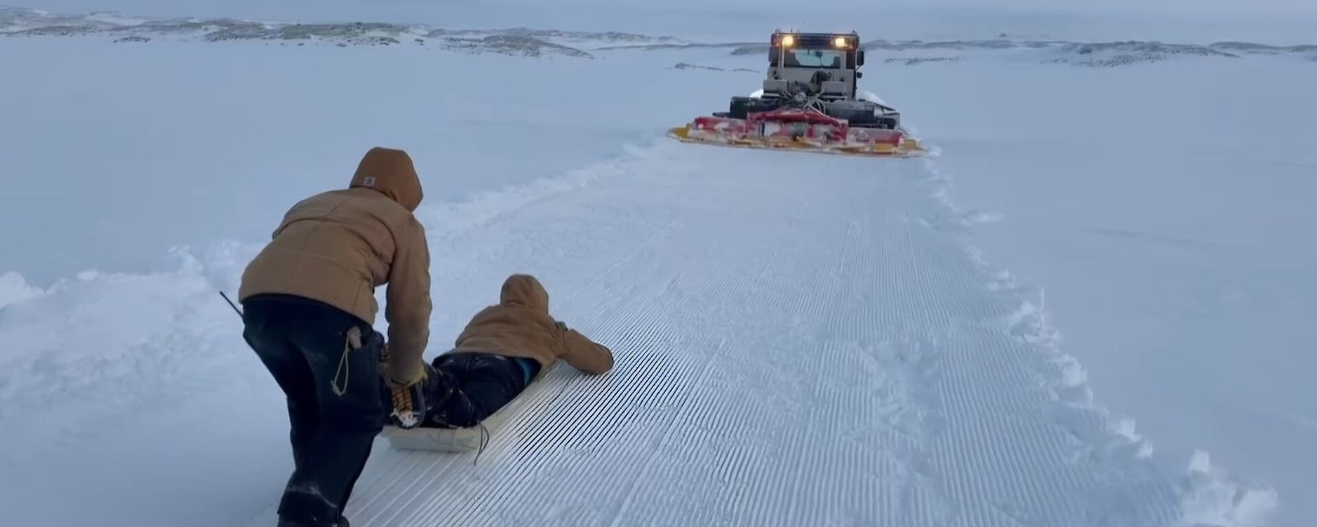 A person lying face down on a sled in the snow with another person pushing them, and a snow groomer with its lights on ahead