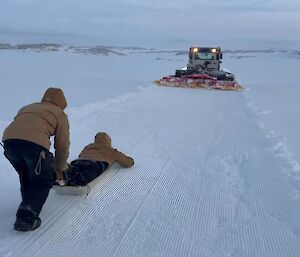 A person lying face down on a sled in the snow with another person pushing them, and a snow groomer with its lights on ahead