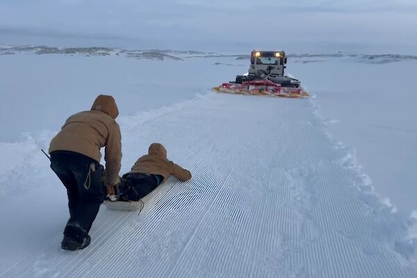 A person lying face down on a sled in the snow with another person pushing them, and a snow groomer with its lights on ahead