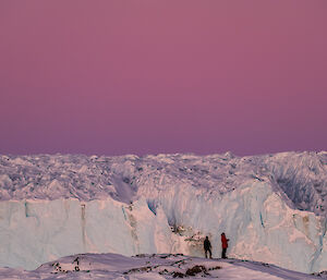 A full moon is visible in a pink sky above a blue glacier, with two expeditioners visible in the foreground