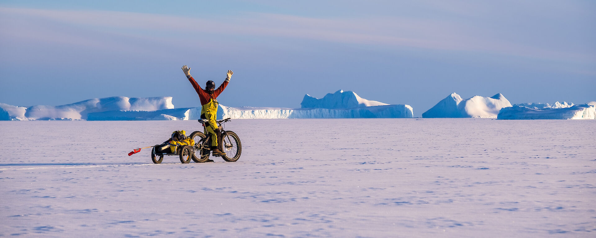 A man on a bicycle towing a small trailer on sea ice raises his arms in triumph