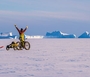 A man on a bicycle towing a small trailer on sea ice raises his arms in triumph