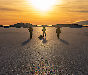 The silhouette of three people walking across sea ice away from camera, dragging sleds, into a golden sunset