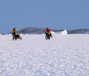 Two expeditioners on bikes ride across sea ice under a blue sky