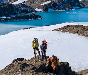 Expeditioners pose on a rock in front of a vivid blue lake with rocky moutains in the background