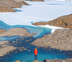An expeditioner wearing an orange backpack surveys a snowy and rocky landscape