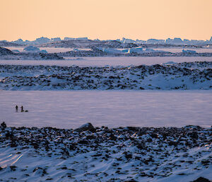 A broad view of sea ice with a golden sunset and the silhouette of two expeditioners