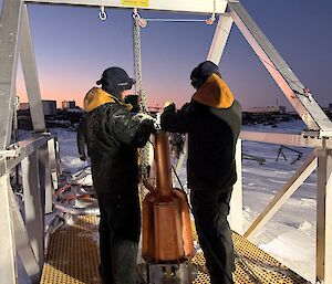Two expeditioners with the station's melt bell - a big bronze-coloured cylinder that gets lowered into a cavern to melt ice for the station's water supply