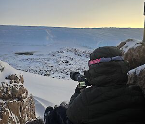 An expeditioner perched on rocks with a camera, taking photos of an emperor colony below