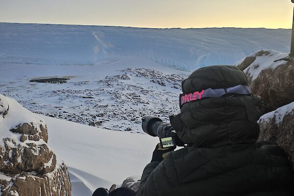 An expeditioner perched on rocks with a camera, taking photos of an emperor colony below