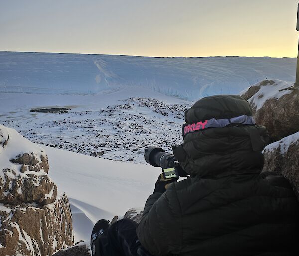 An expeditioner perched on rocks with a camera, taking photos of an emperor colony below