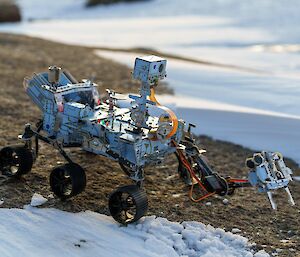 A model Mars Rover seemingly taking samples via a mechanical arm, on a rocky outcrop with sea ice in the background