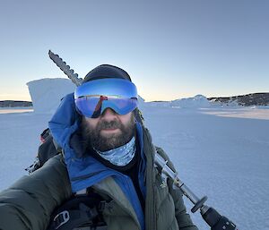 An expeditioner on the sea ice, carrying a sea ice drill - with iceberg and rocky cliffs in the background