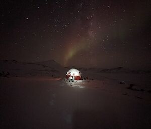 A clear picture of an apple hut, with light showing through round windows under a starry night sky with a green and red aurora visible, positioned as if it is coming out of the hut