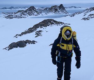 Expeditioner wearing black and yellow walking through rocky and snowy terrain.