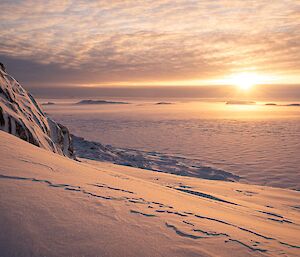 a snowy mountain stands in front of the ocean with the sun setting in the distance