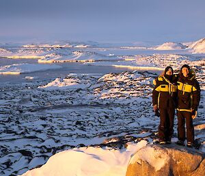 Two expeditioners stand on top of a rocky peak above the snowy landscape