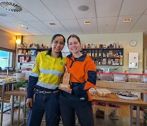 An expeditioner in yellow high visibility clothing presents a trophy to another expeditioner in an orange high visibility jumper