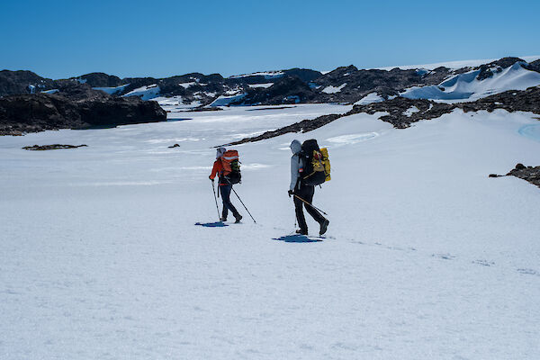 Two people walking across a snowy plain with black mountains in the background