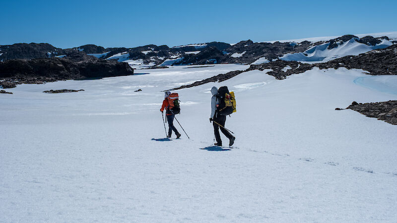 Two people walking across a snowy plain with black mountains in the background