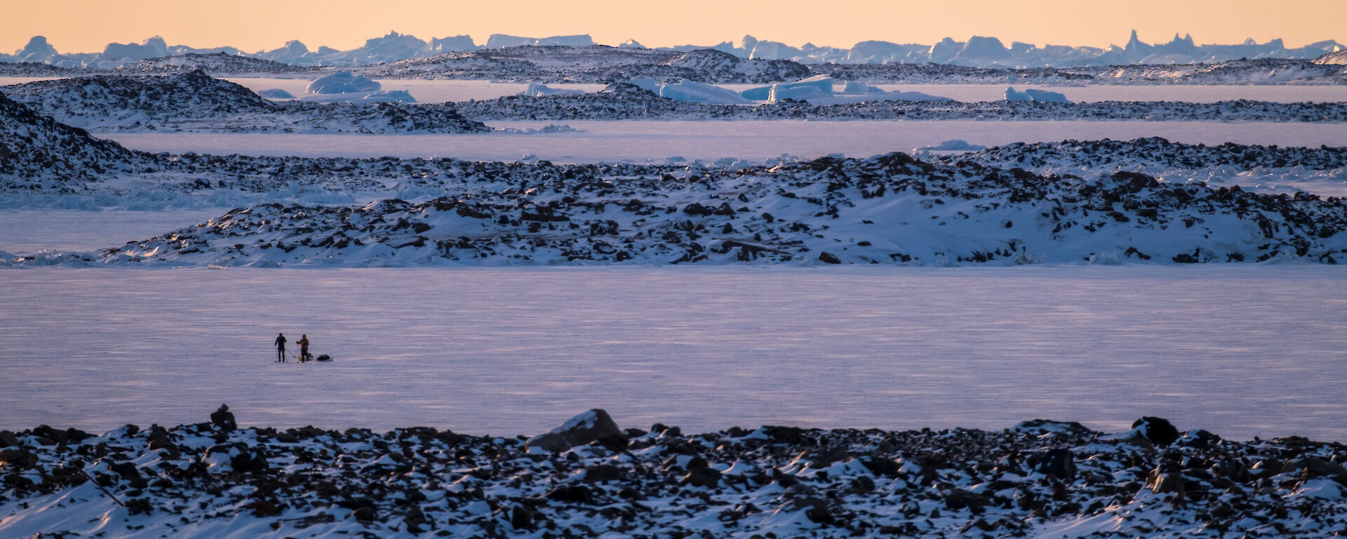 Two people in the distance walking on a snowy plain with icebergs in the distance