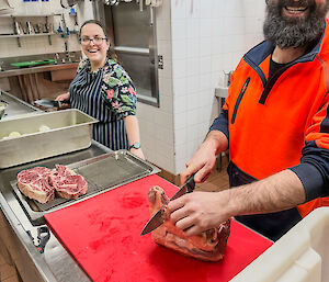 A man stands in a kitchen smiling and cutting up meat