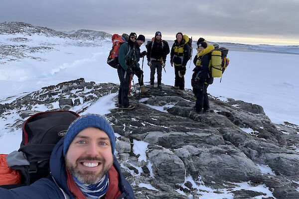 A man takes a selfie of himself and five others on some rocks with snow all around them