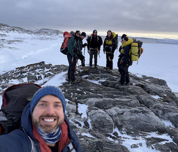 A man takes a selfie of himself and five others on some rocks with snow all around them