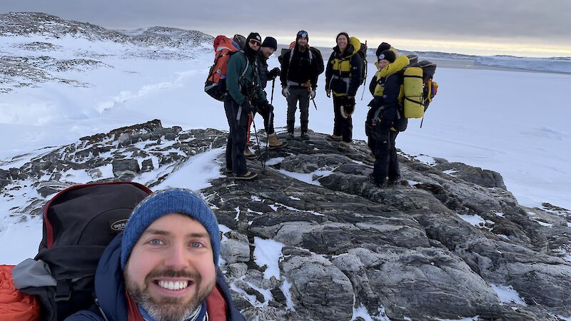 A man takes a selfie of himself and five others on some rocks with snow all around them