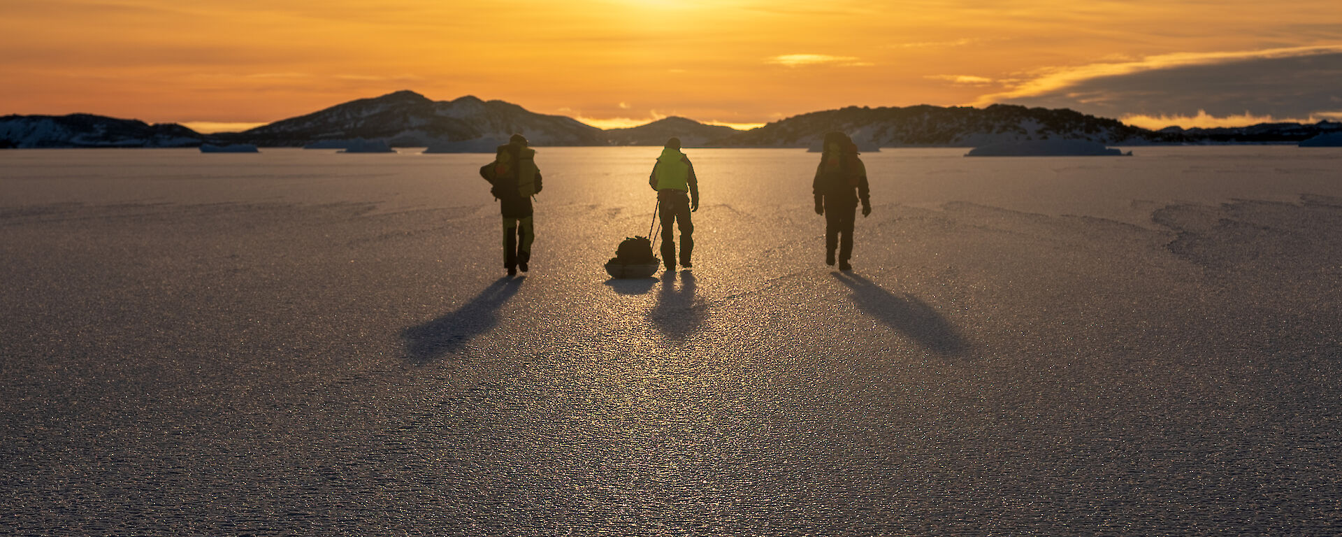 Rear shot of three people walking across the ice with golden sunshine in the distance