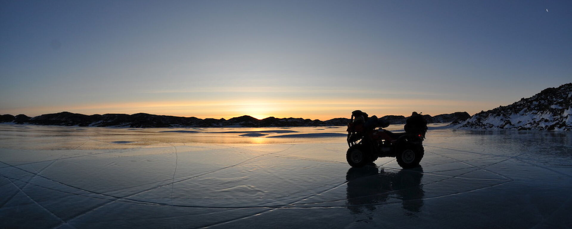 a quad bike in shadows on the ice, as the sun sets below the horizon