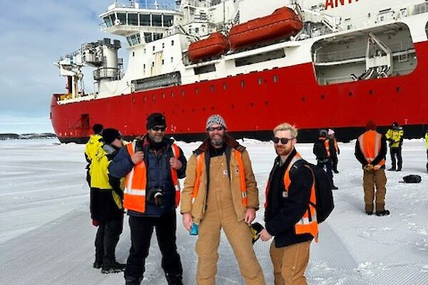 Three people in high visibility vests pose on the sea ice infront of a large red ship