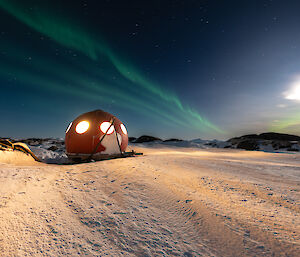 A green aurora trail and bright moon shines over Macey apple field hut