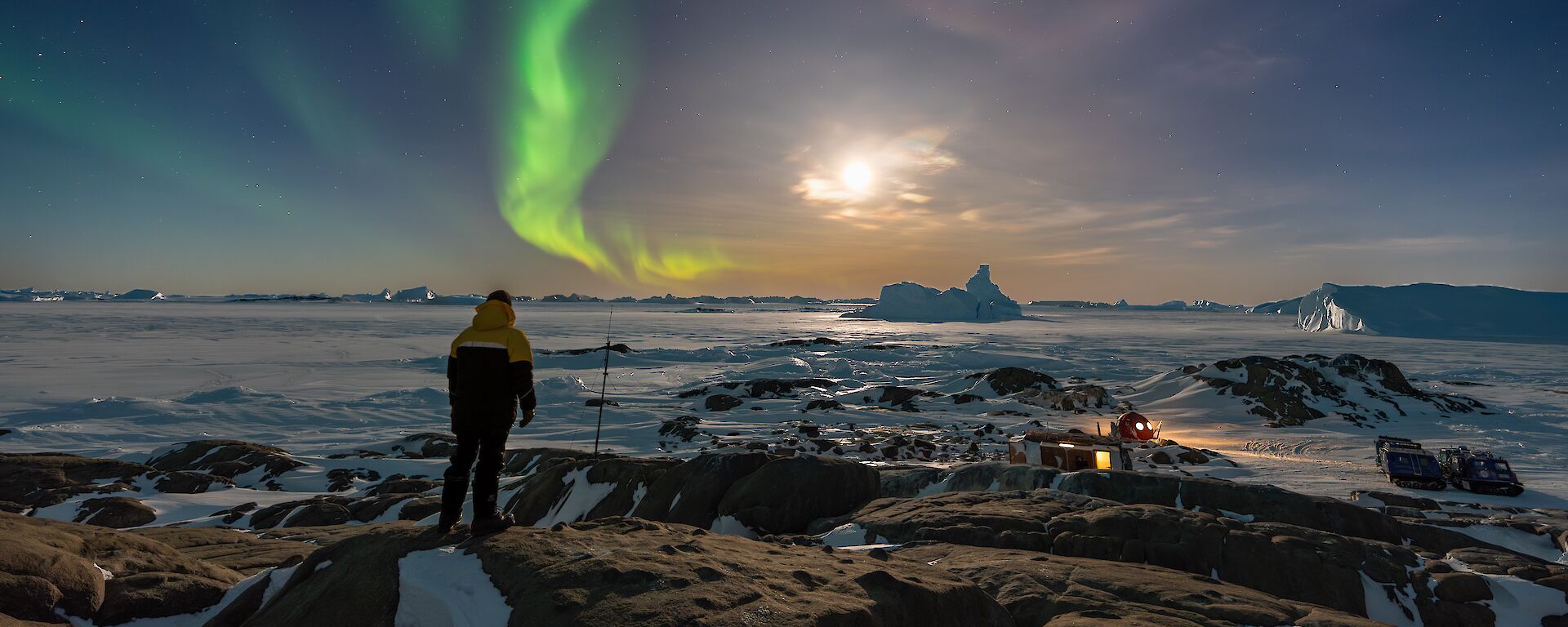 An expeditioner standing on a hill with green aurora trail and bright moon lighting up the camp area and sea ice