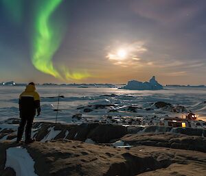 An expeditioner standing on a hill with green aurora trail and bright moon lighting up the camp area and sea ice