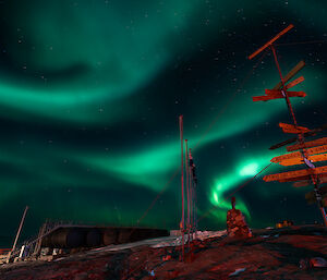 Mawson's flagpoles and sign lit up with overhead green aurora trails in the sky