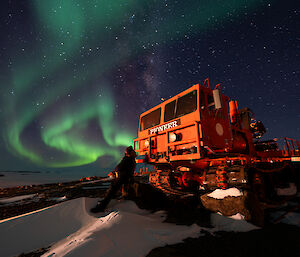 Sweeping aurora in sky with expeditioner leaning on an old pioneer tractor in foreground