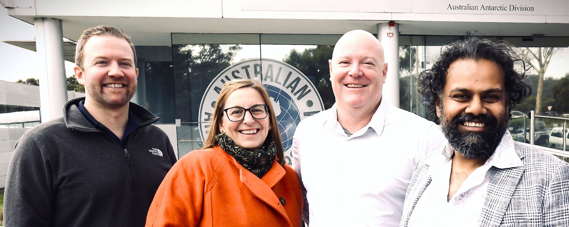 3 men and one woman stand together in front of a white and glass building. They are smiling.