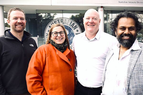 3 men and one woman stand together in front of a white and glass building. They are smiling.