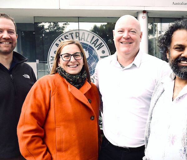 3 men and one woman stand together in front of a white and glass building. They are smiling.
