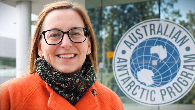 A woman wearing black-framed reading glasses, and orange overcoat and a dark coloured scarf; stand in front of a building with the logo for the Australian Antarctic Program prominently displayed.