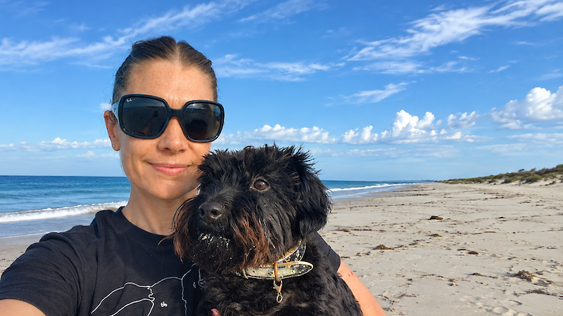 A women wearing large, black sunglasses is holding a black, fluffy dog in her arms. Behing them is a long stretch of beach under a blue sky.