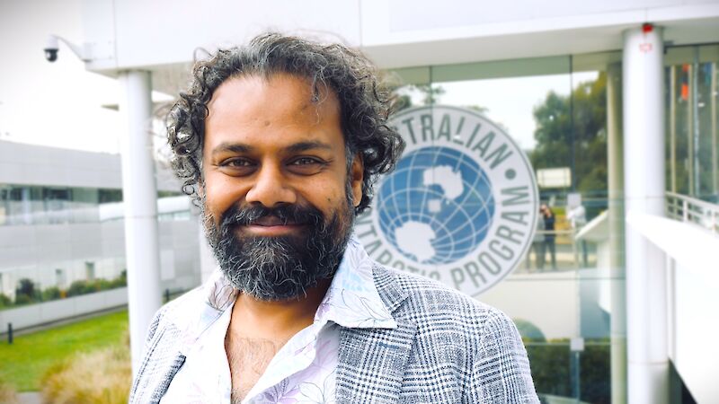 A man wearing a white business shirt and checked overcoat stands smiling, in front of a building with the logo for the Australian Antarctic Program prominently displayed.