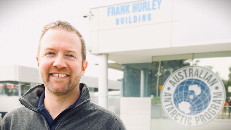 A man wearing a black, collared sweater, stands smiling, in front of a building with the logo for the Australian Antarctic Program prominently displayed.