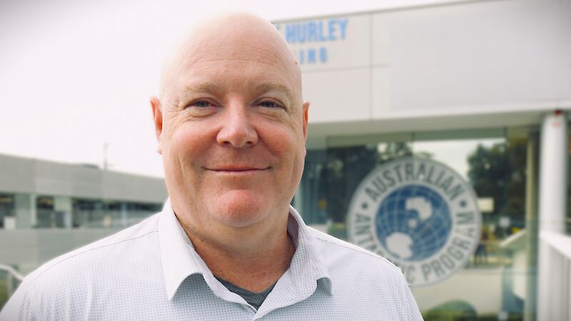 A man wearing a white business shirt stands smiling, in front of a building with the logo for the Australian Antarctic Program prominently displayed.